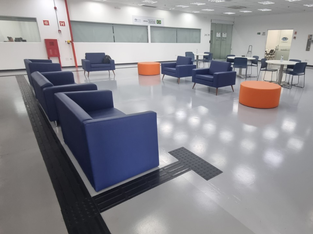 Sala corporativa com piso epóxi cinza claro, mobiliário azul e laranja, e faixa tátil preta para acessibilidade.