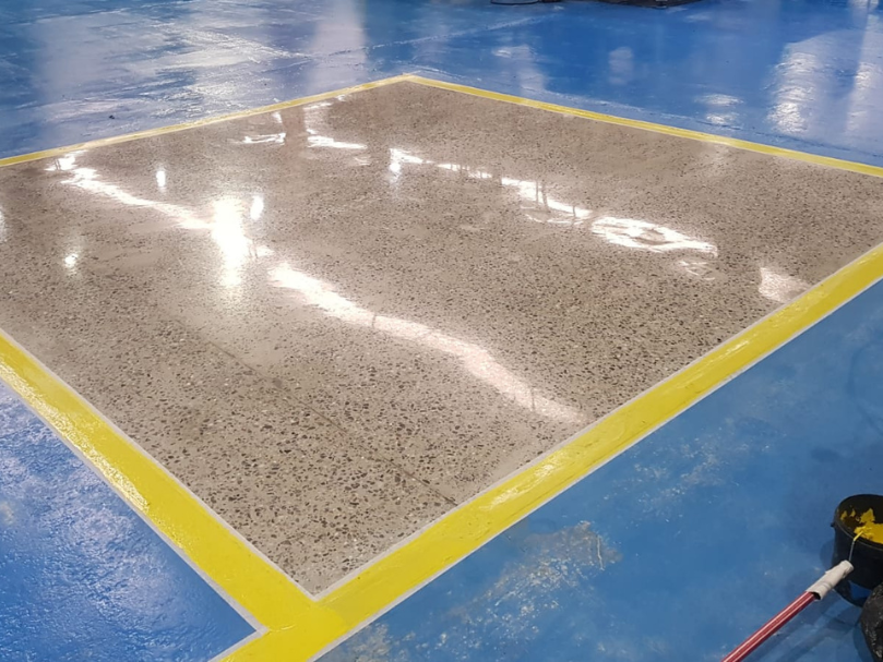 Quadro de piso polido delimitado por faixas amarelas sobre piso epóxi azul, utilizado como área demonstrativa.