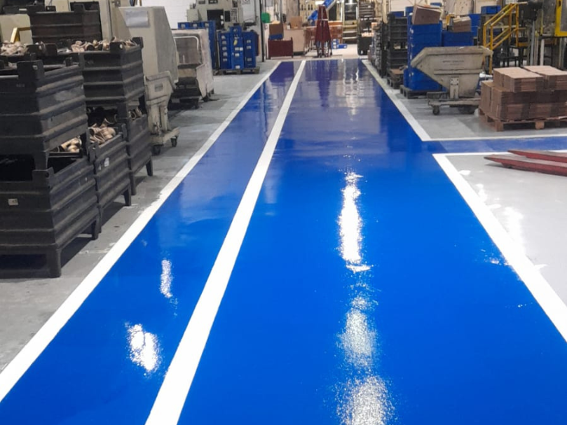 Corredor industrial com piso epóxi azul brilhante, demarcado por faixas brancas, em área de produção com paletes e máquinas ao redor.