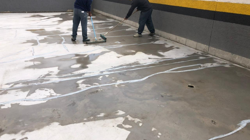 Dois homens usando rodos para espalhar material sobre um piso de concreto parcialmente molhado, com várias áreas remendadas e marcas de infiltração.