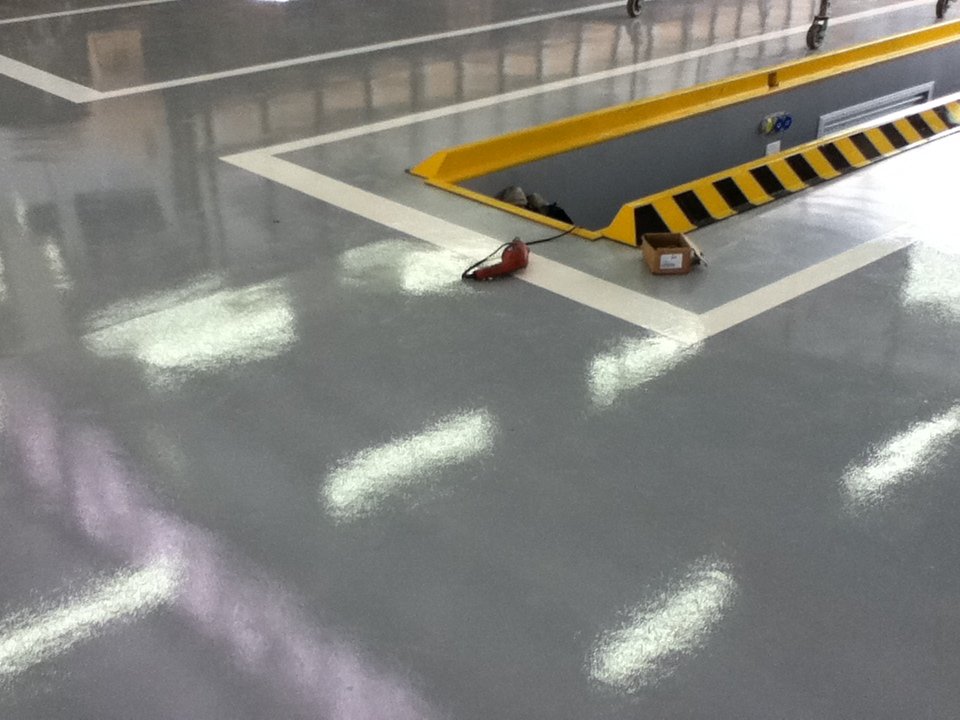 Piso epóxi cinza brilhante ao redor de valeta de manutenção, com demarcações brancas e áreas de segurança em amarelo e preto.