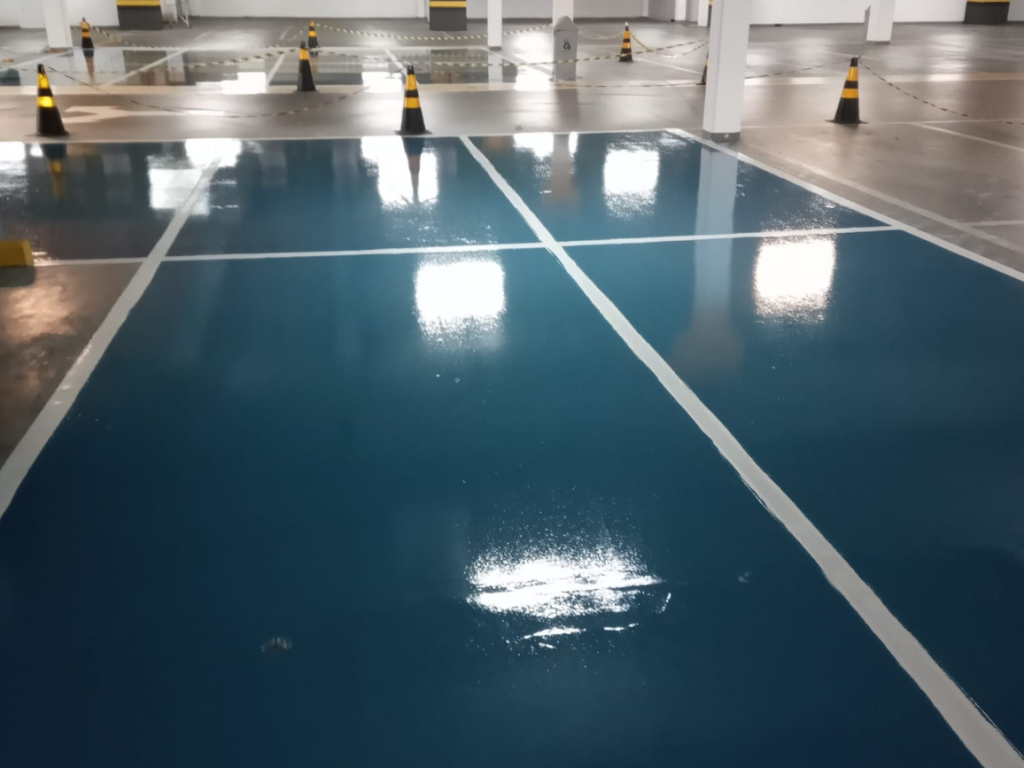 Piso epóxi azul brilhante recém-instalado em estacionamento coberto, com demarcações brancas e cones de isolamento.