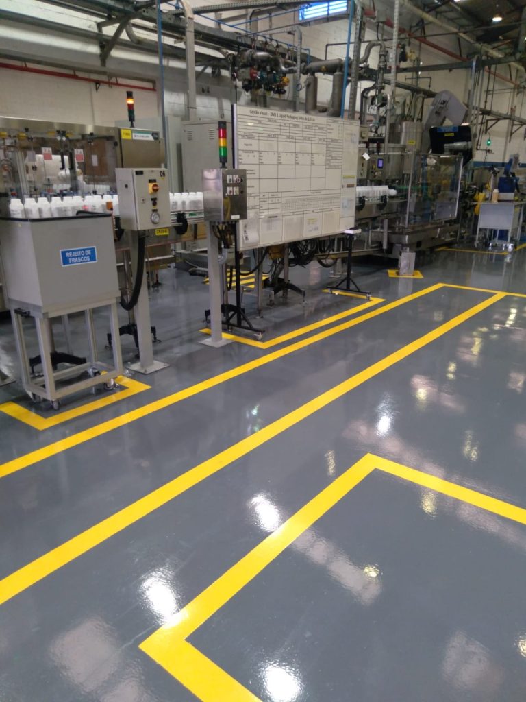 Piso epóxi cinza brilhante em área industrial, com demarcações amarelas bem definidas em volta de máquinas e painéis.