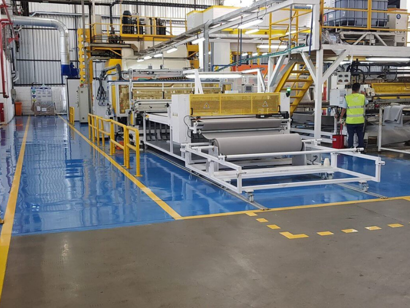 Linha de produção industrial com piso epóxi azul brilhante e demarcações amarelas, máquinas e operador trabalhando ao fundo.