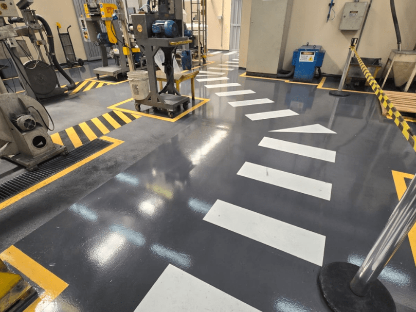 Corredor industrial com piso epóxi cinza escuro e setas brancas indicando direção, cercado por máquinas e barreiras de segurança.