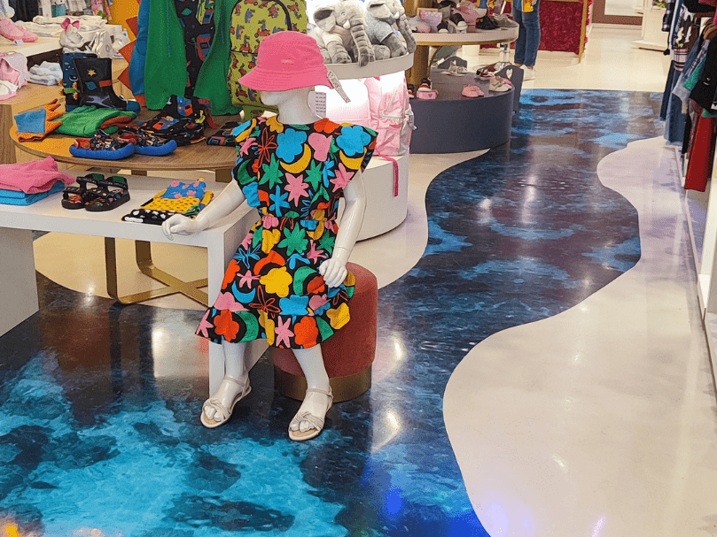 Corredor de loja infantil com piso epóxi azul marmorizado e áreas em branco, com manequim vestindo roupas coloridas.