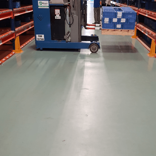 Equipamento industrial em operação sobre piso epóxi verde claro, em corredor de estoque com prateleiras metálicas.