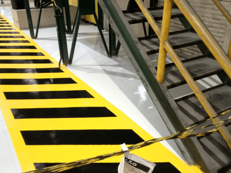 Área industrial com piso epóxi branco e faixa de segurança amarelo-preto aplicada ao lado da escada metálica.