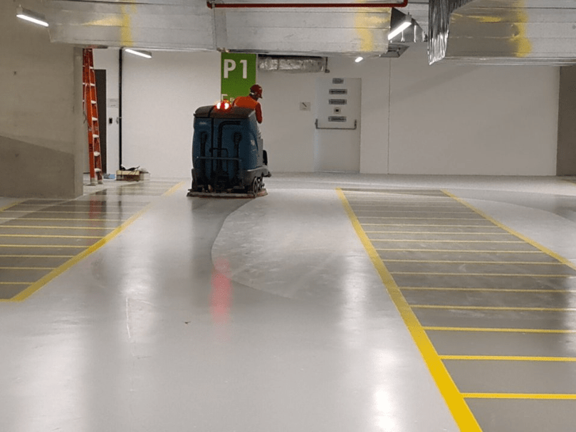 Funcionário operando máquina de limpeza industrial sobre piso epóxi cinza, com marcações amarelas de estacionamento.