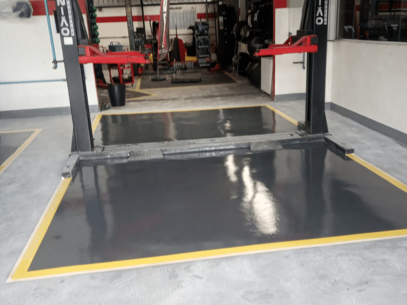 Área de elevador automotivo com piso epóxi preto brilhante, demarcado com faixa amarela em ambiente de oficina.