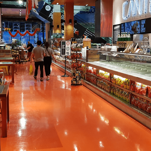 Corredor de supermercado com piso epóxi laranja brilhante, clientes caminhando e vitrines de alimentos ao lado.