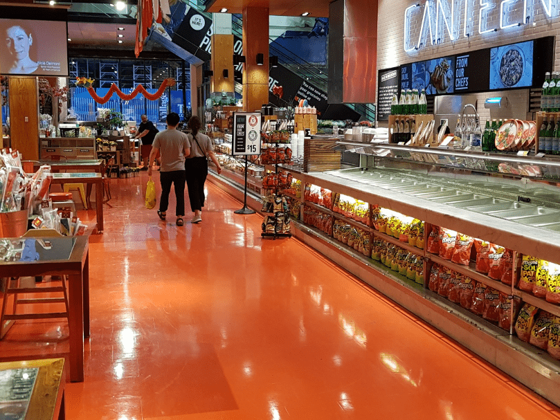 Supermercado com piso epóxi laranja brilhante, expositores de alimentos e clientes caminhando pelo corredor principal.