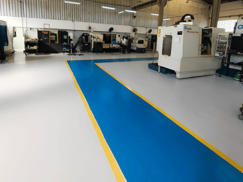 Área industrial com piso epóxi branco e faixa azul demarcando circulação, máquinas CNC distribuídas ao fundo.