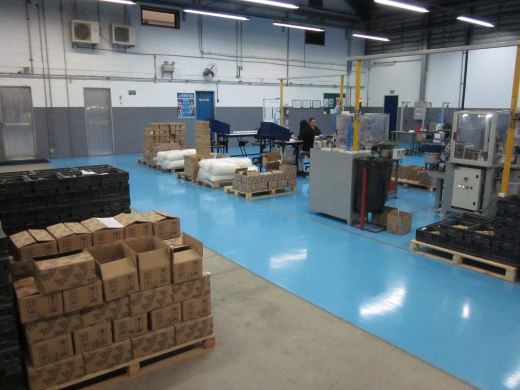 Galpão de produção com piso epóxi azul brilhante, paletes com materiais e máquinas industriais distribuídas pelo ambiente.