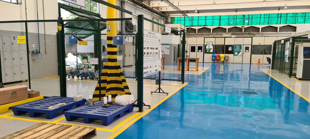 Galpão industrial com piso epóxi azul e cinza brilhante, demarcações amarelas e estruturas metálicas ao redor da área de trabalho.