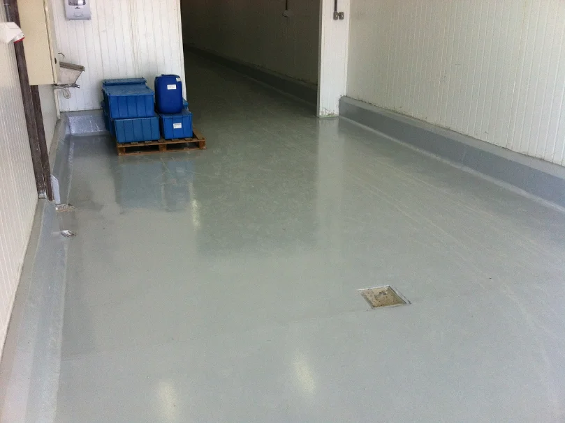 Corredor industrial com piso autonivelante cinza claro, caixas plásticas armazenadas em pallet ao fundo e ralos no chão.