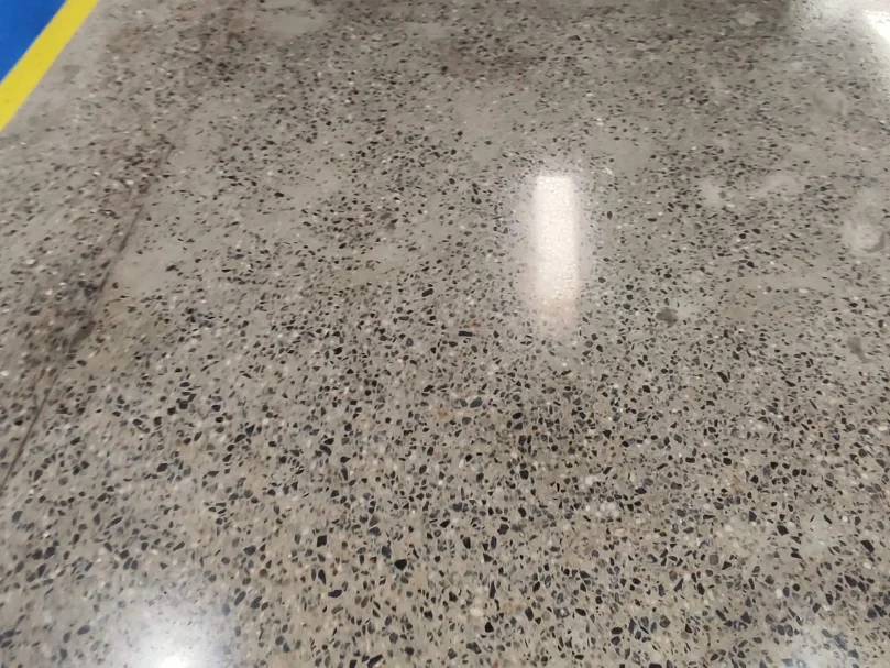 Piso de concreto polido com agregados expostos e superfície brilhante próxima a faixa de sinalização amarela.