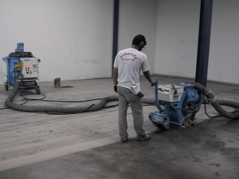 Técnico operando máquina de jateamento abrasivo para preparar concreto antes da aplicação de novo revestimento.