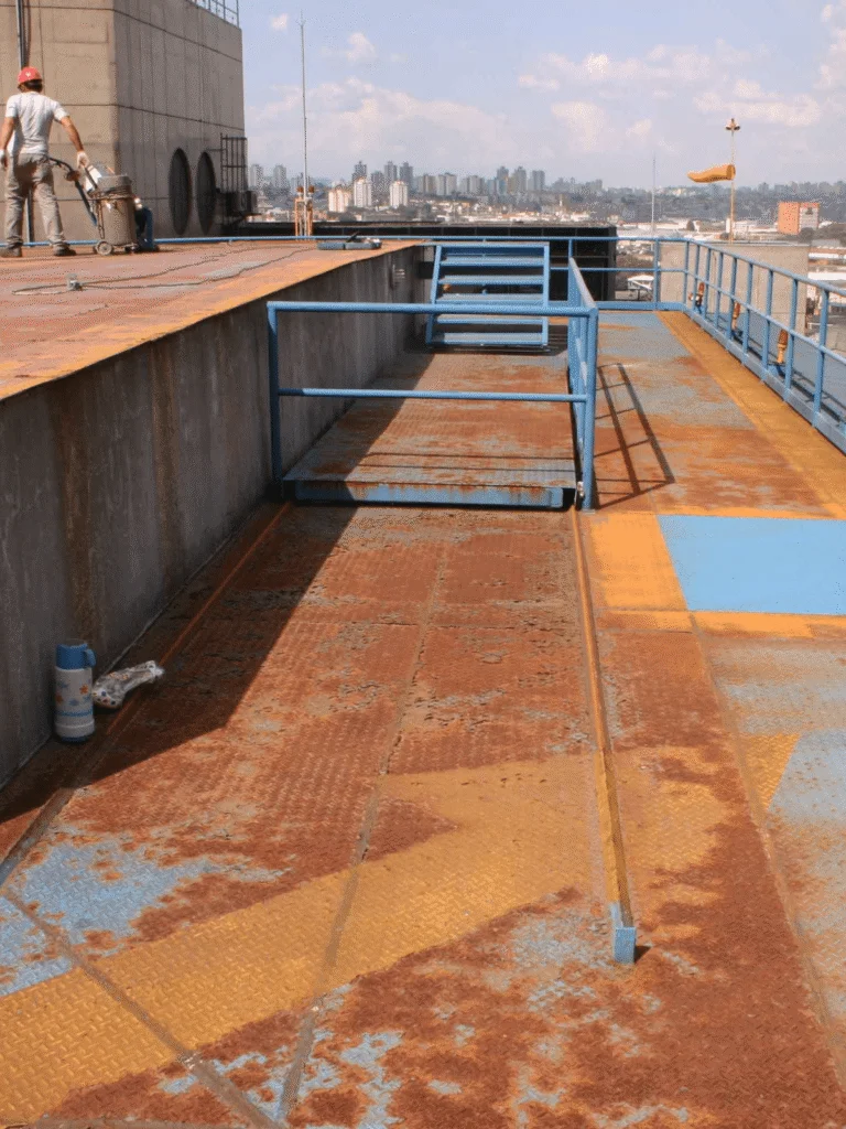 Superfície de heliponto desgastada com áreas de ferrugem e pintura deteriorada, enquanto trabalhador realiza manutenção no local.