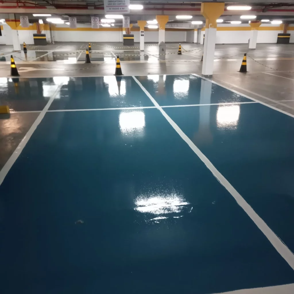 Piso epóxi azul brilhante recém-aplicado em estacionamento coberto, com demarcações brancas e cones de sinalização ao fundo.