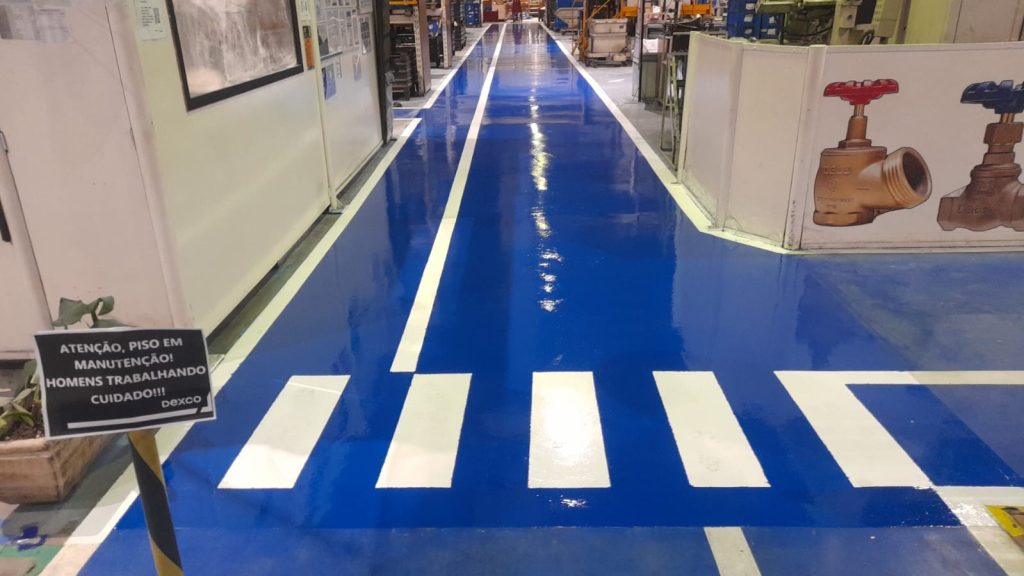 Corredor industrial recebendo pintura epóxi azul com faixas brancas para demarcação de segurança.