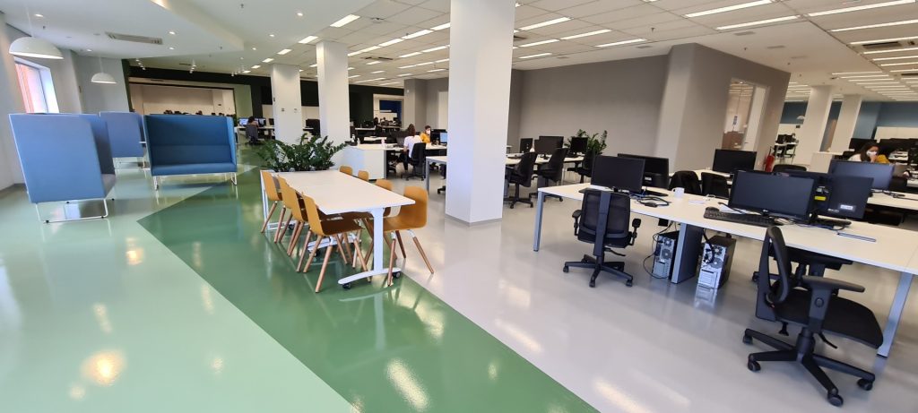 Área ampla com piso epóxi verde em diferentes tons, mesas de convivência e sofás azuis.