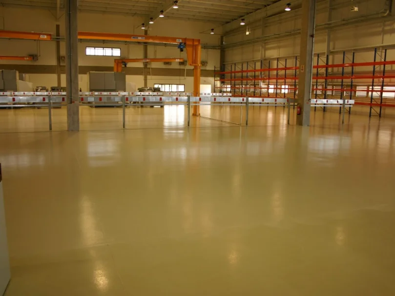 Galpão industrial amplo com piso resinando claro recém-instalado, iluminação superior e estruturas metálicas ao fundo.