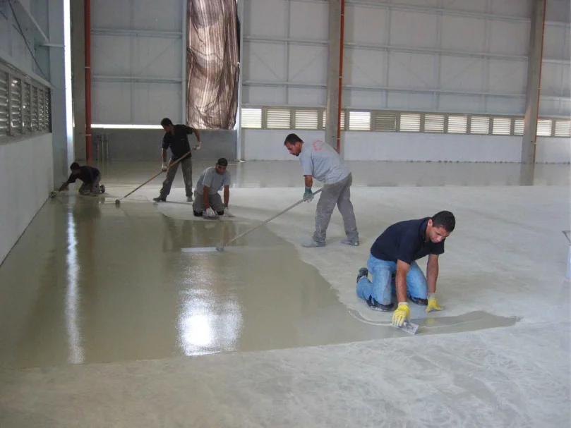 Operário limpando ou preparando o piso com um rodo após aplicação de primer escuro, em uma área ampla de galpão.