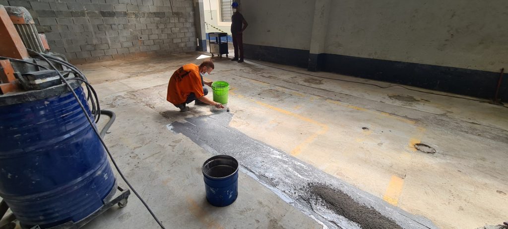 Trabalhador aplicando revestimento epóxi em trecho danificado de piso industrial usando rolo e balde verde.
