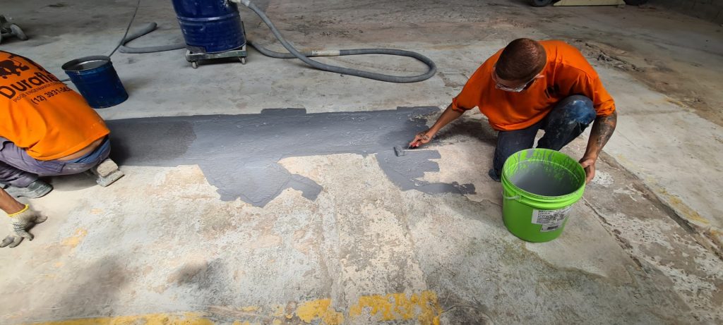 Piso industrial sendo reparado com aplicação manual de argamassa epóxi em área desgastada.