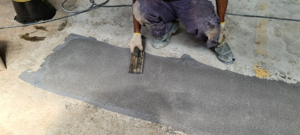 Aplicação manual de revestimento antiderrapante sobre concreto, com trabalhador usando desempenadeira para nivelar o material granulado.