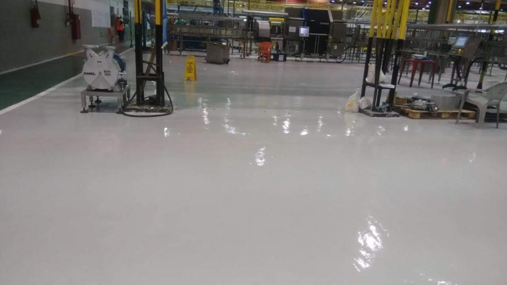 Piso industrial com acabamento brilhante recém-executado, localizado em área fabril com equipamentos e sinalização de segurança.