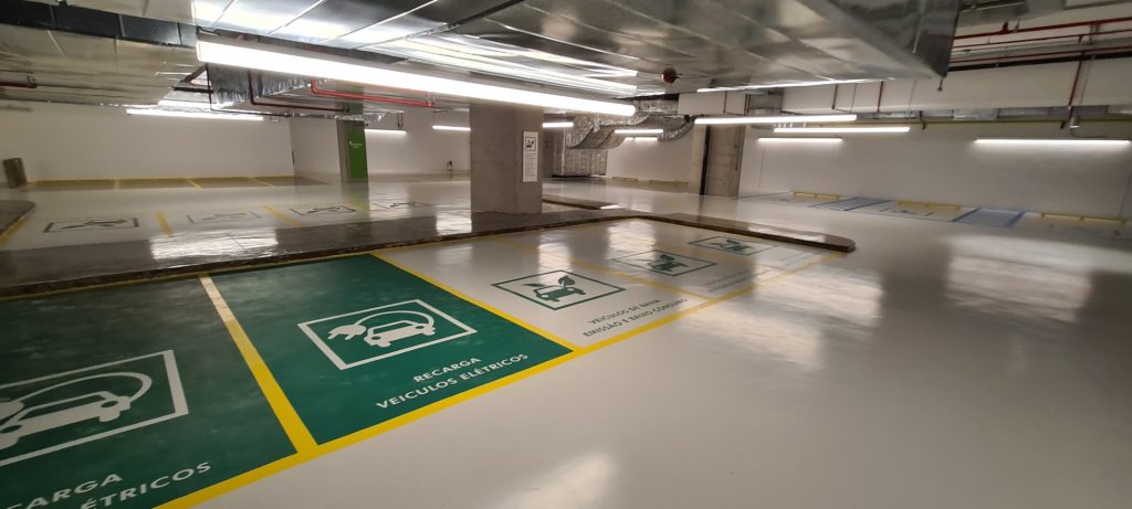 Área de estacionamento para veículos elétricos com piso epóxi verde e branco, com ícones de recarga e faixas amarelas.
