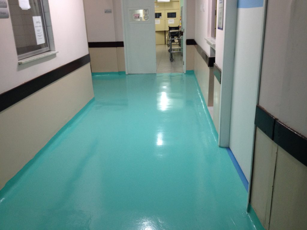 Corredor de hospital com piso epóxi azul brilhante, paredes claras e portas ao fundo, transmitindo ambiente limpo e higienizado.