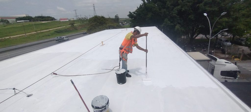 Trabalhador utilizando rolo de pintura para aplicar revestimento branco refletivo sobre um telhado industrial, usando EPIs e preso a cabo de segurança.