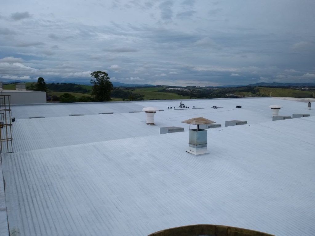 Vista panorâmica de telhado industrial impermeabilizado em branco com chaminés e paisagem urbana ao fundo.