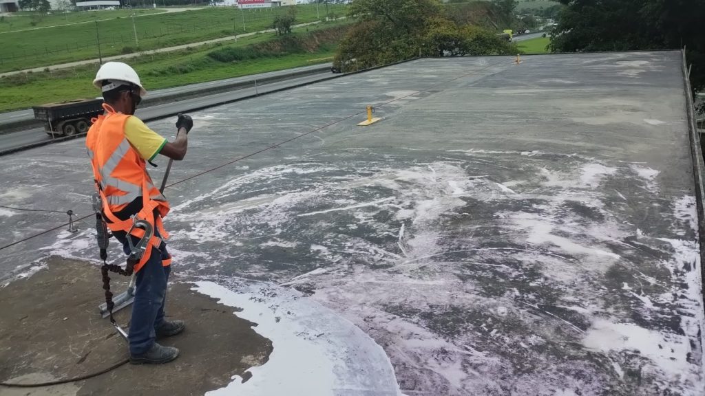 Trabalhador limpando e preparando a superfície suja e escurecida do telhado industrial para aplicação do revestimento.