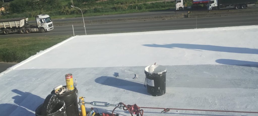 Baldes de tinta e equipamentos de aplicação sobre o telhado em processo de pintura branca, com tráfego de caminhões ao fundo.