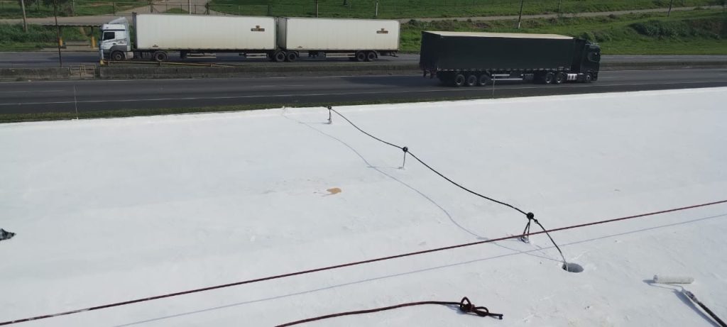 Telhado industrial recém-pintado com revestimento branco, com amarrações de segurança e caminhões passando na rodovia ao fundo.
