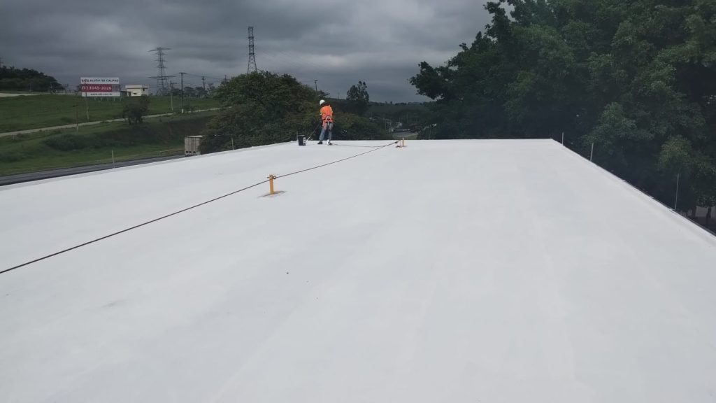 Vista ampla de um telhado industrial totalmente coberto por revestimento branco refletivo, com trabalhador finalizando o serviço ao fundo.