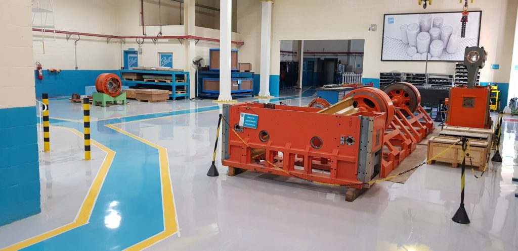 Área industrial com piso epóxi cinza e faixas demarcatórias amarelas e azuis, máquinas laranjas e equipamentos ao fundo.