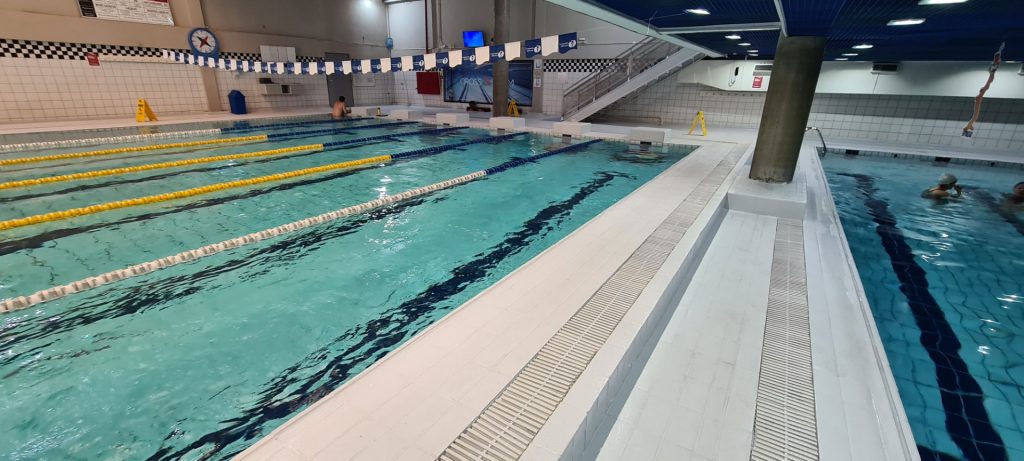 Piscina semiolímpica com raias amarelas e azuis, bordas brancas e área molhada revestida com piso antiderrapante claro.
