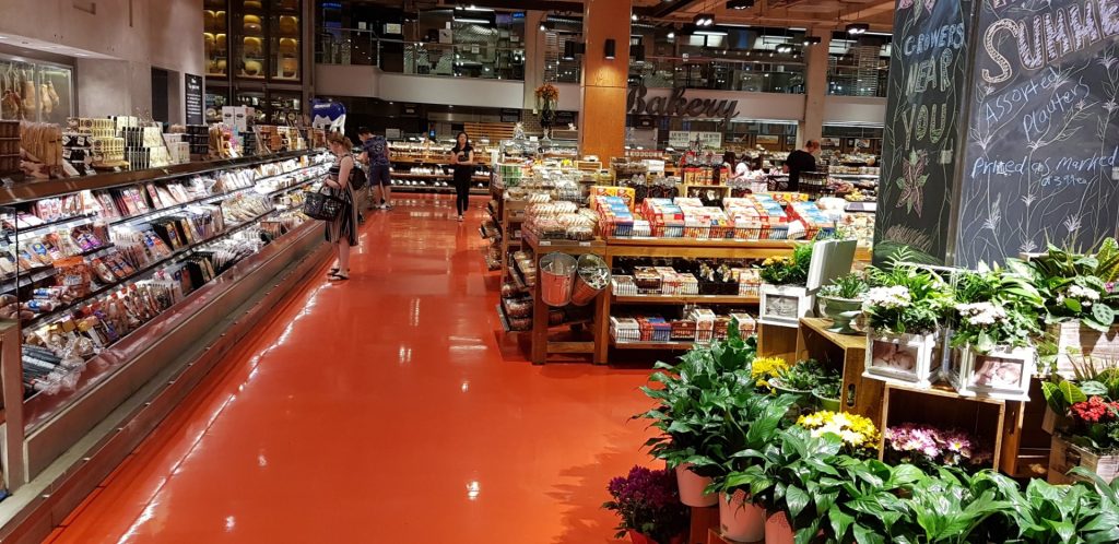 Área de frios e padaria com piso epóxi vermelho brilhante, gôndolas repletas de produtos e clientes circulando.